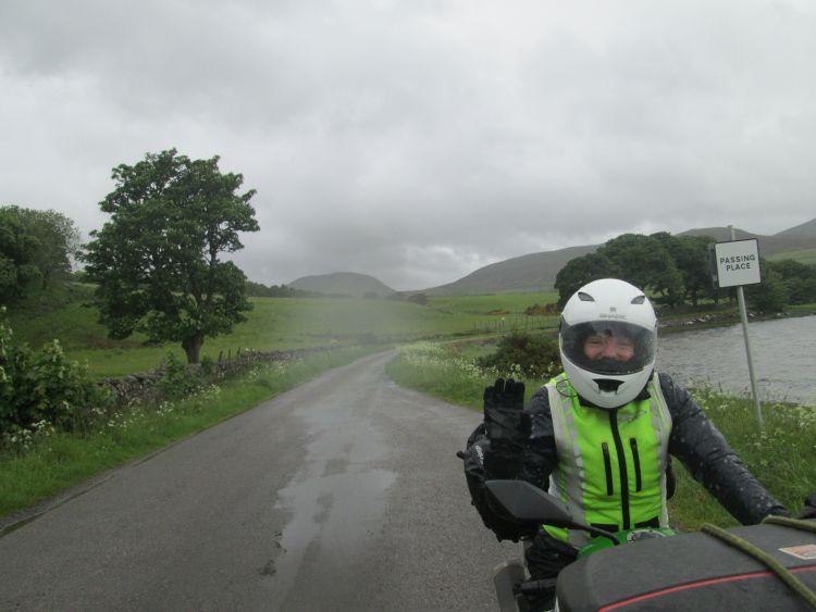 Sharon waves at the camera from her bike in dark skies and rain falls all around
