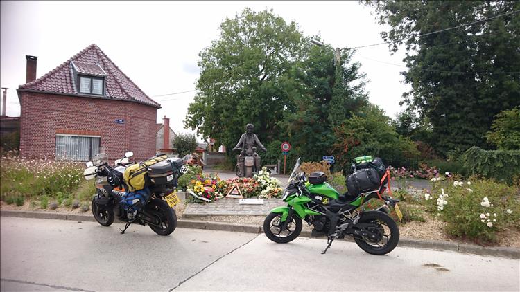 A statue featuring a world war 2 motorcyclist surrounded by flowers and the small village