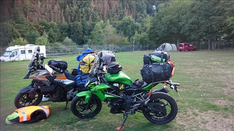 The 2 motorcycles on a field and a tree lined hill behind