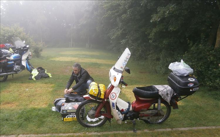 A rider is packing his camping gear onto a Honda C90 in the misty murky fog of the campsite
