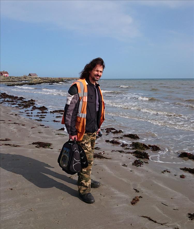 Ren stands in his bike gear with the wind and sun on his face at the beach in Ballywalter