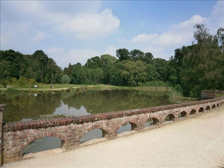 A low ornate brick wall in front of a large pond and lush green trees at the pretty park in Passchendaele