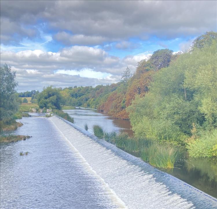 A broad slow flowing river tips over a small weir in luscious tree filled countryside