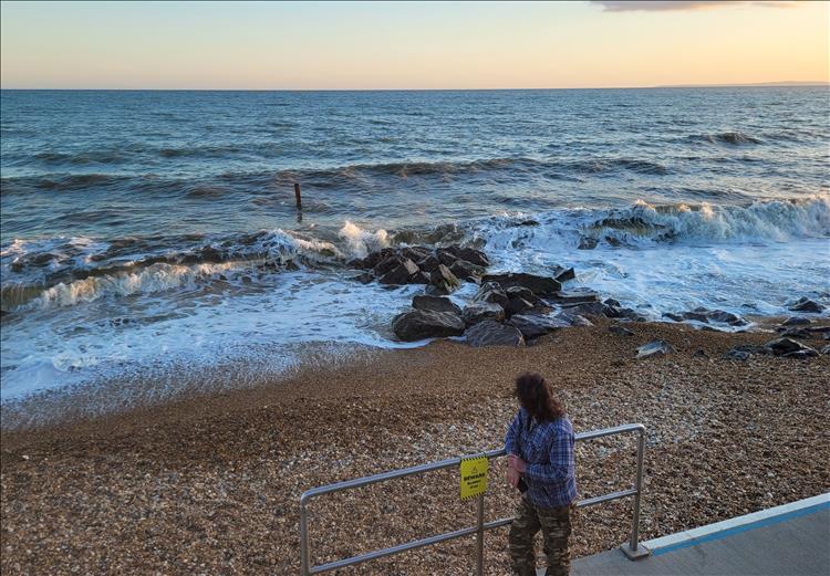 Ren stands against a railing on the sea defences looking over a shingle beach and out to sea