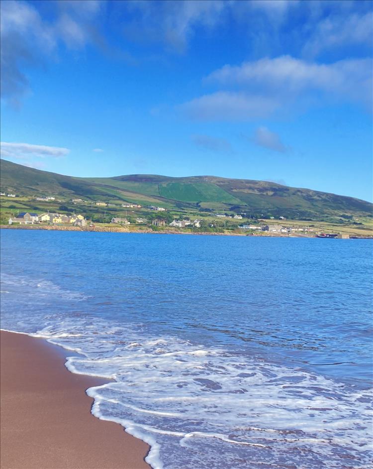 Gentle waves lap a sandy beach with hills and a small town in the distance