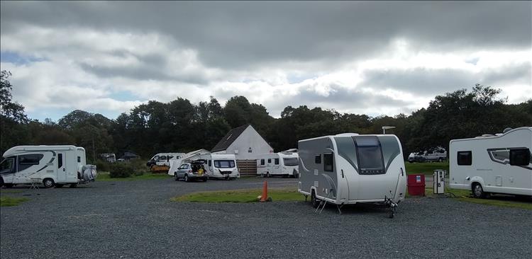 Gravel hardstanding, some grass, various buildings, and a line of trees at the campsite