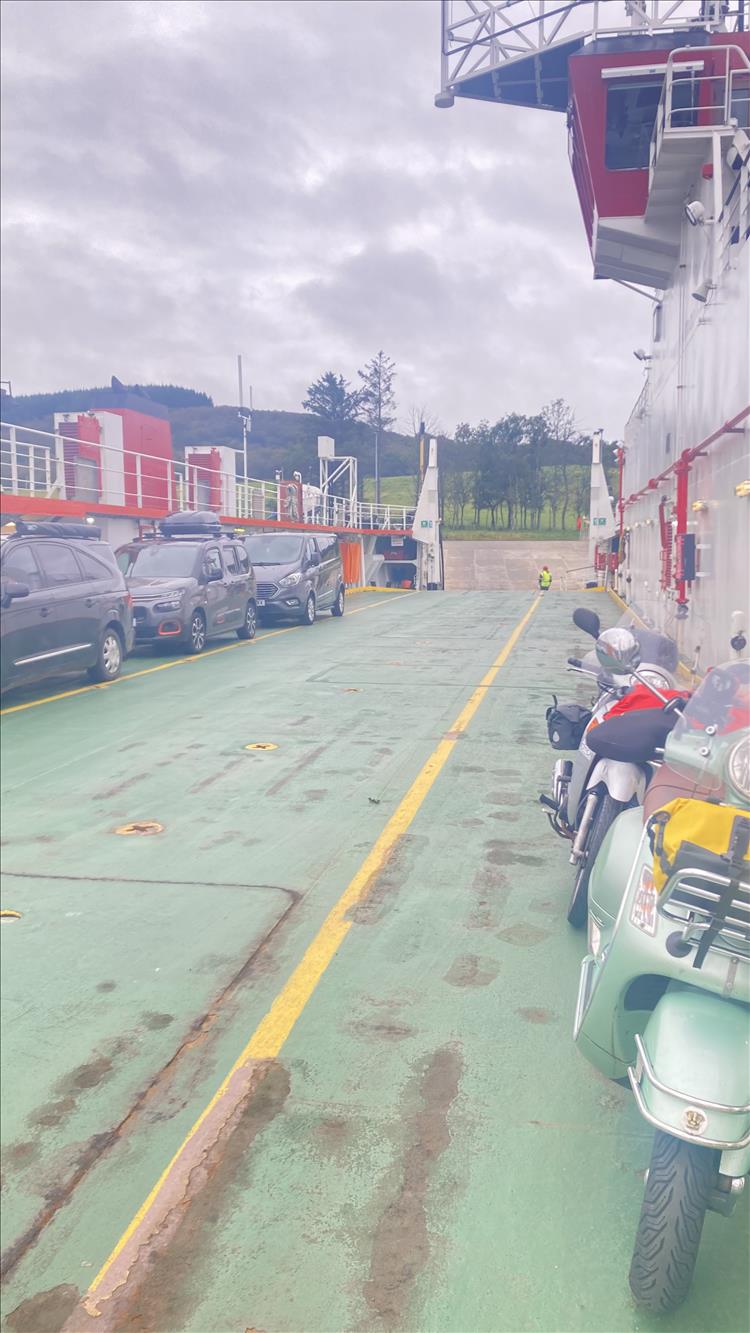 We see the wide deck of the ferry, the scoots parked on the deck and the scenery behind the deck