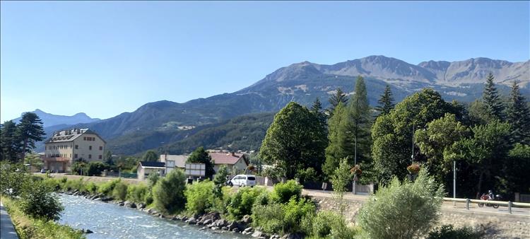 Clear hazy blue skies, towering mountains and a pretty river seen in The Alps
