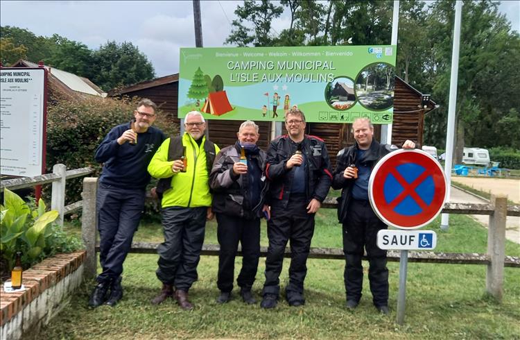 The friends stand by the camping municipal sign all smiling and enjoying a drink