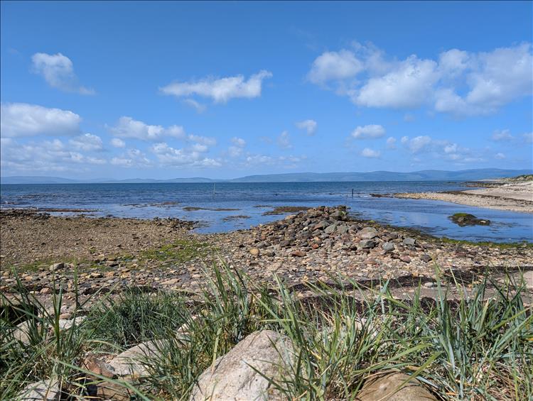 Looking over a sonty shore we see waters, distant mountains and blue skies with some light cloud
