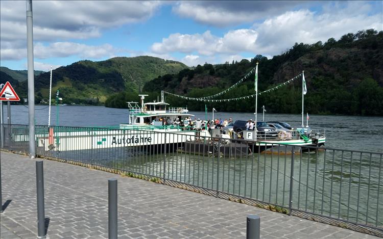 A small flat top ferry with a few cars and passengers, thickly wooded hills on the other side of the river