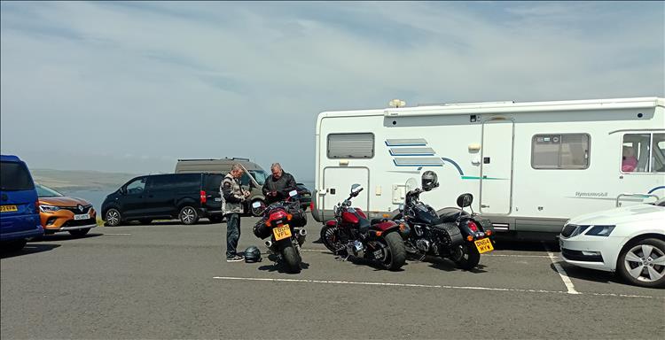 Ren's friends and their bikes parked by a massive motorhome and various cars near the lighthouse
