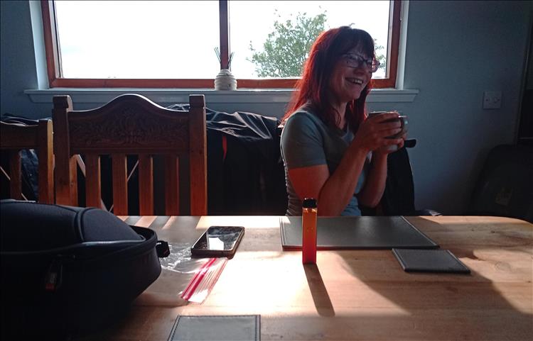 Sharon laughing and smiling with a brew, oddly lit with a window behind her