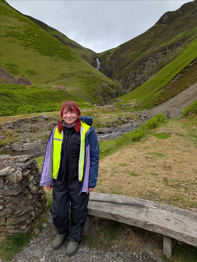 Sharon in her waterproofs and bike gear smiles, behind are steep hills and a narrow waterfall