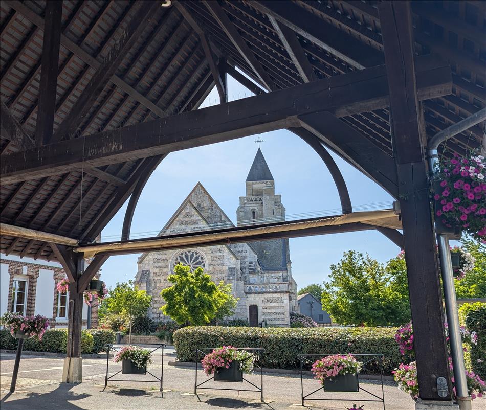 From under a tall wooden open walled structure we see a church. There's flowers and trees and neatly trimmed bushes