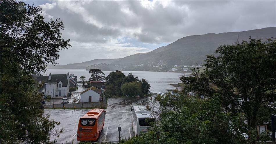 Looking from a hillside down to a car park, a vast Loch, and mountains in the rainy distance