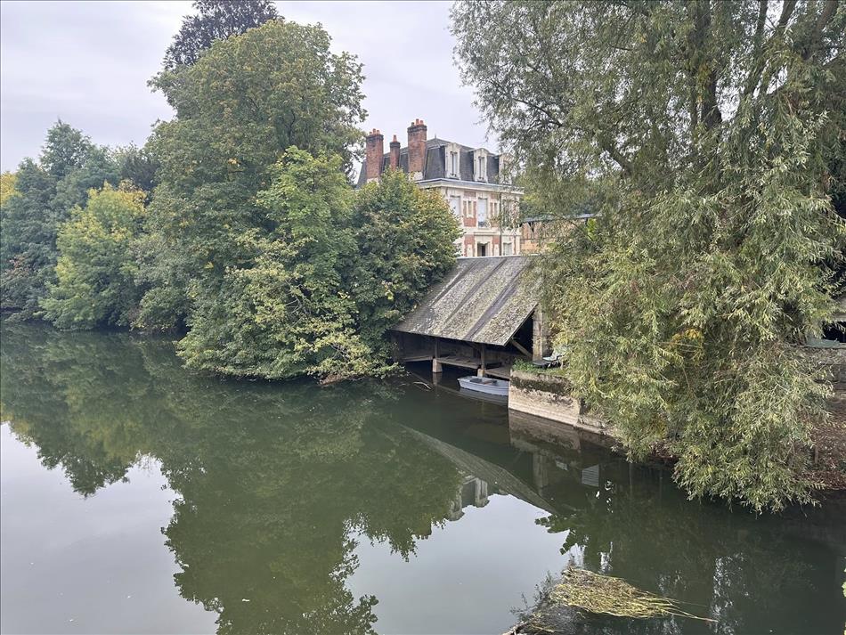 A calm river with a small boat, trees aplenty and between the tree French houses