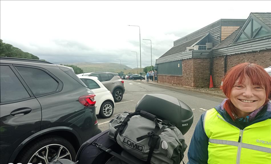 A big smile from Sharon next to her bike and cars in the car park. We can make out misty mountains of The Lake District