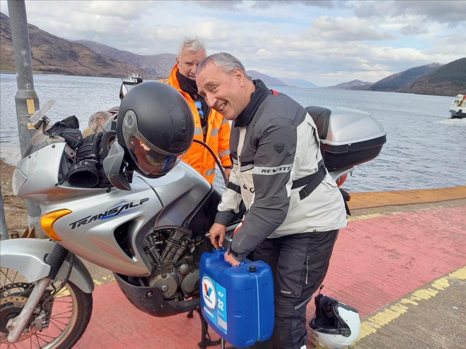 A chap smiles while holding a large oil container and a pipe, they're siphoning the diesel from the tank