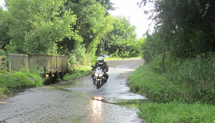 A motorcycle and rider splash through a small ford on a leafy lane