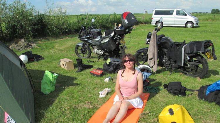 Sharon sits on the grass surrounded by the camping gear, smiling in the sun