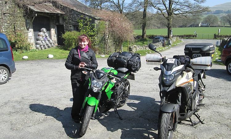 Sharon stands next to both motorcycles in the sun outside the countryside pub
