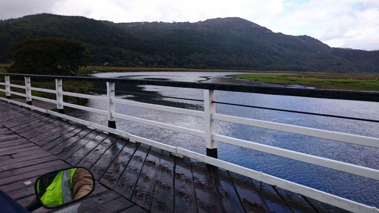 Looking out from the wooden bridge there are trees covering hills and the broad river. Grey skies