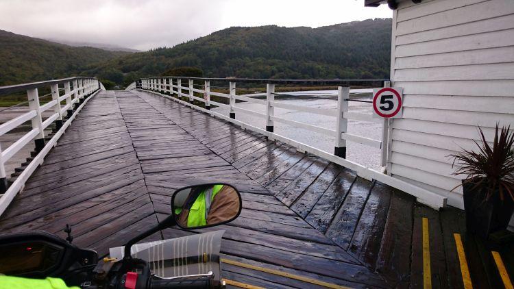 A long old wooden bridge across a broad river, trees cover misty mountains in the background