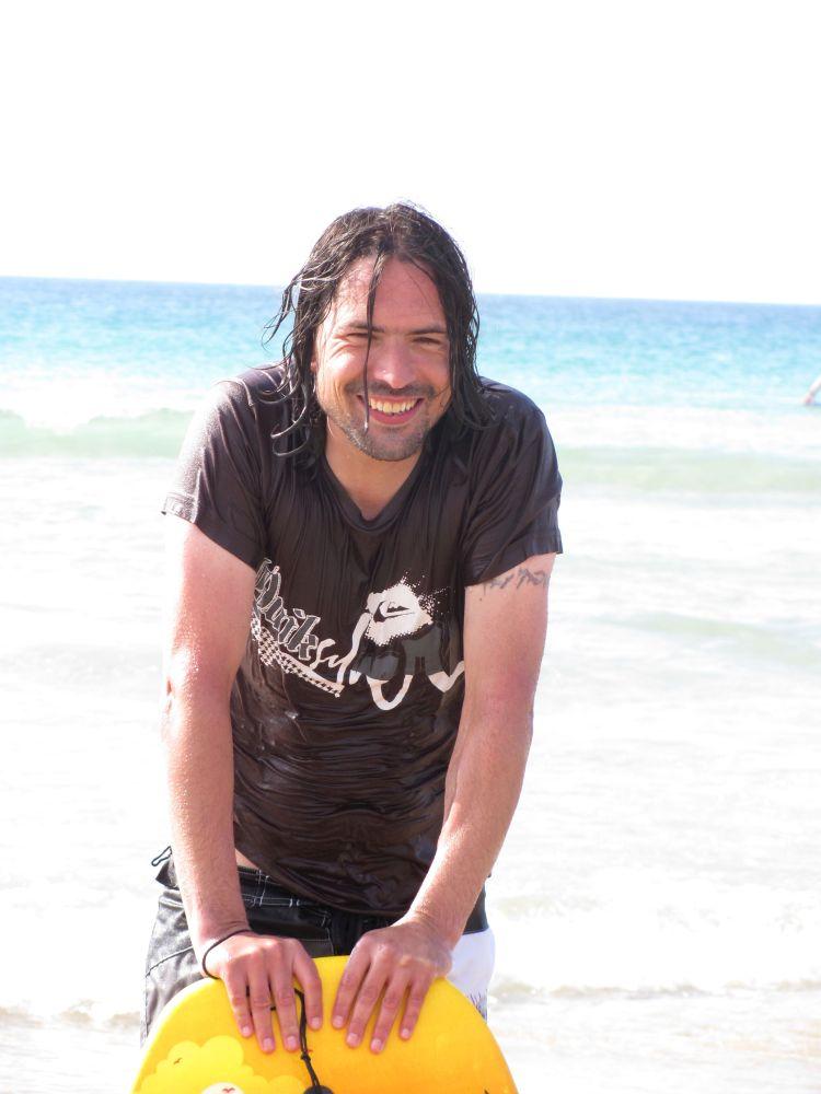 Ren leans against a bodyboard, all wet and smiling while in the sun at Cornwall in the past