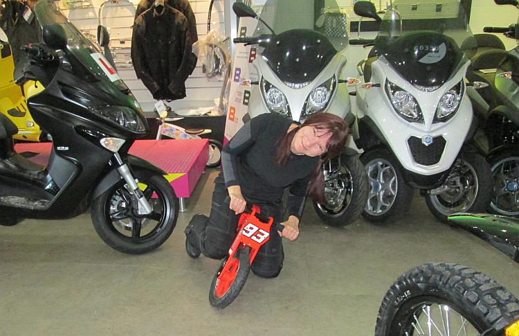 Sharon's on a tiny kids walking-bike, pretending to get her knee down