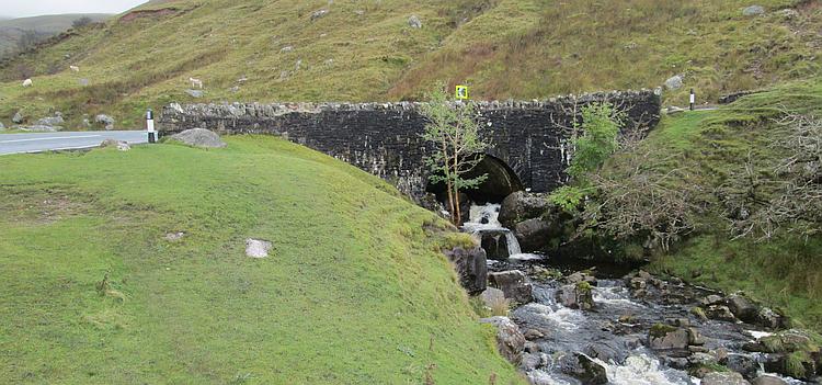 A small bridge over a brook set in among hills covered in hardy grasses and boulders