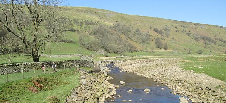 A small river flows through boulders, rolling green hills and glorious blue skies at Swaledale