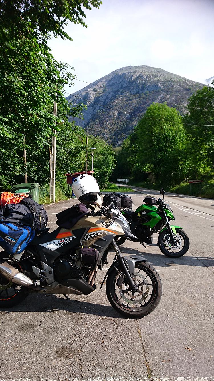 The two motorcycles at Puente De Vage. A large green hill rises in the background of the tiny vilalge