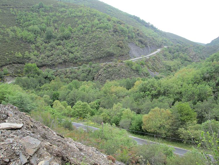 A road cuts along the side of a steep hill covered in hardy trees and scrub