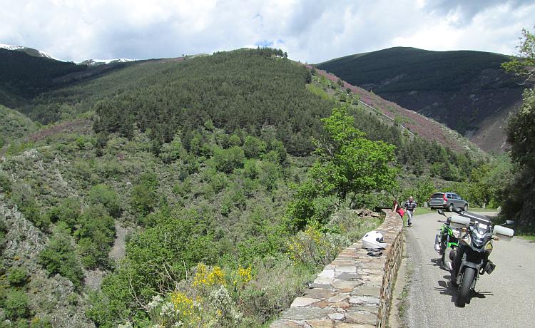 The motorcycle next to a wall looking into and up from a steep tree filled valley