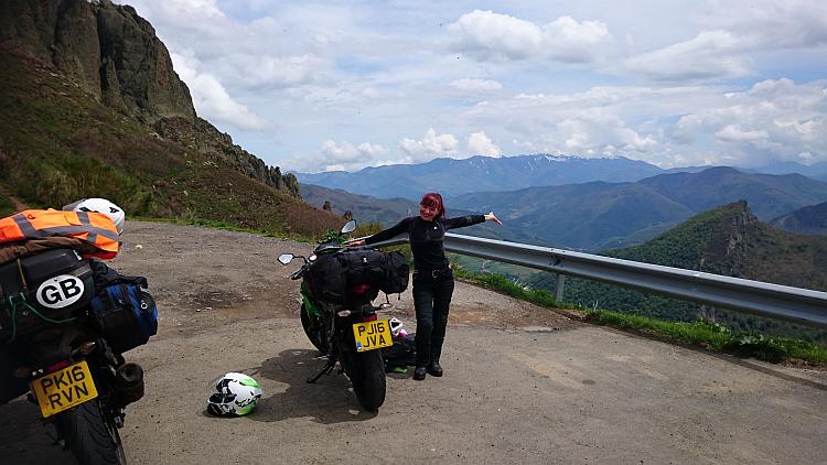 Sharon is smiling with the bikes, arms open presenting a majestic mountain range fading off into the distance
