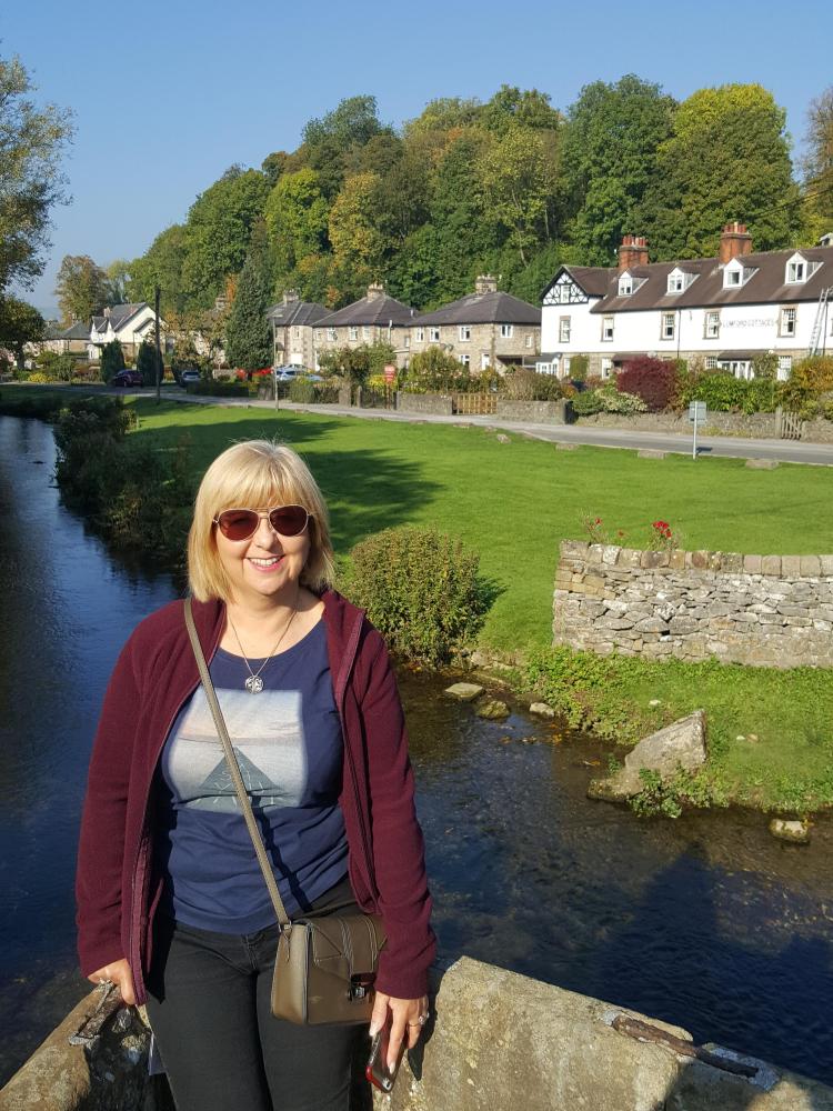 Paula dressed in light walking gear smiling by a pretty river on a sunny day