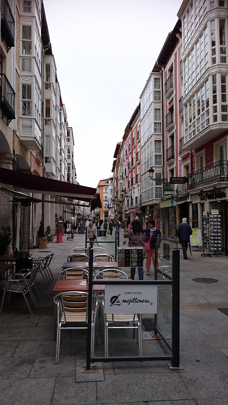 A long street with tall buildings either side, in the foreground chairs from the cafe spill into the street
