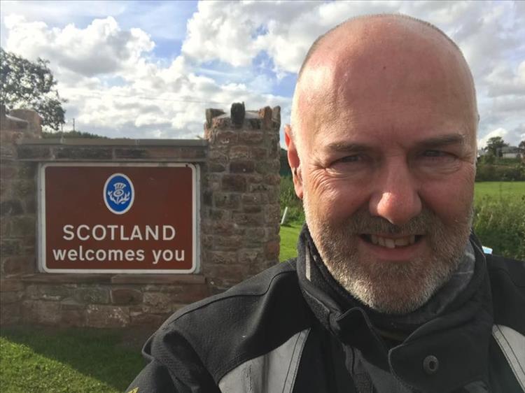Andy smiles standing in front of a brown "Scotland welcomes you" sign at Gretna Green