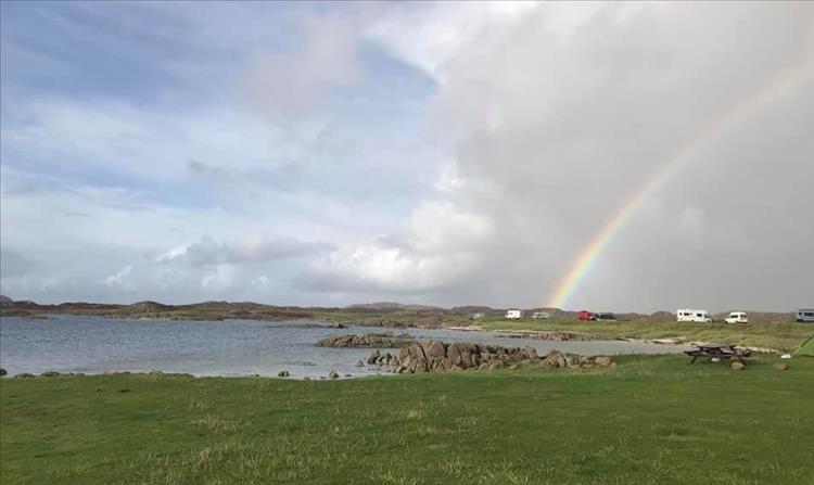 A rainbow drops into the field with campervans and tents by the shoreline at the campsite