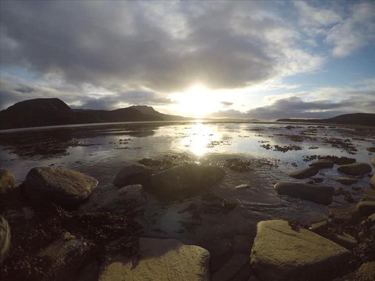 The sun is low in the sky reflecting off the water with rocks and seaweed and mountains