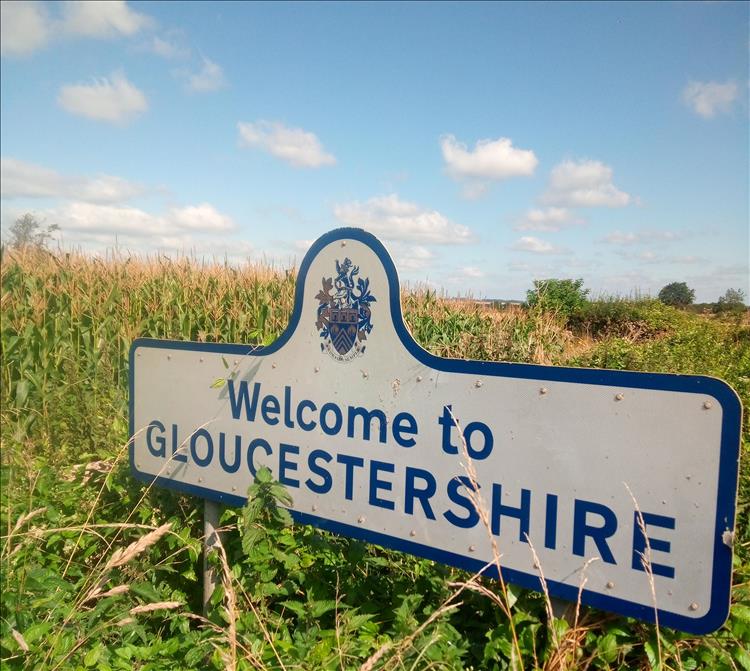 A sign says Welcome To Gloucestershire by the road in the countryside on a sunny day