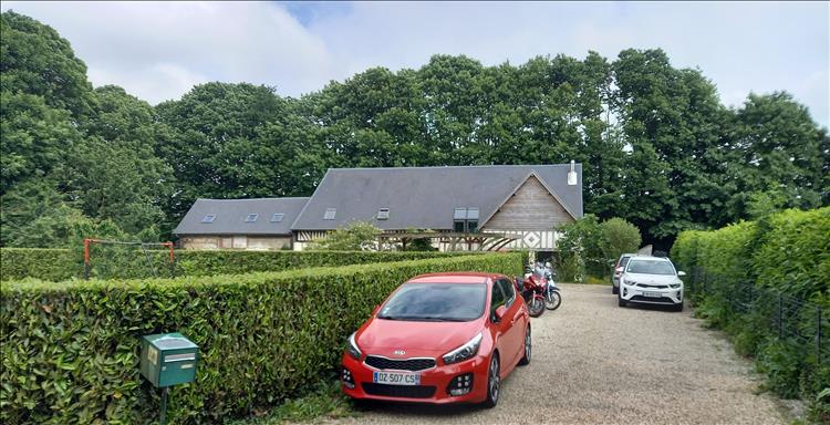 An old wooden frame house set among trees in France. Cars and bikes are in the driveway