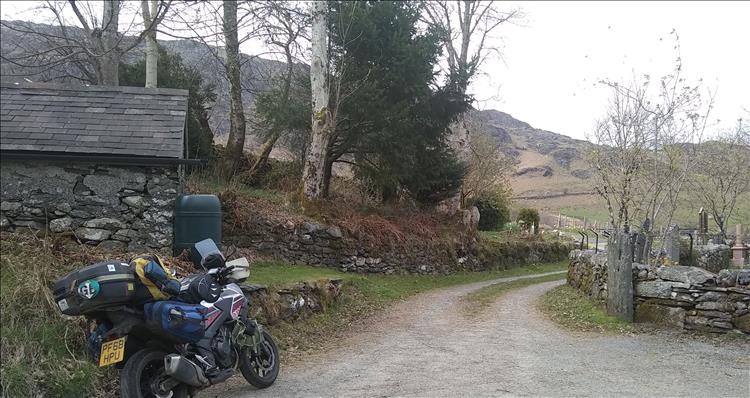 The bike is in the scene of scant old buildings, trees, hardy grasses and mountains