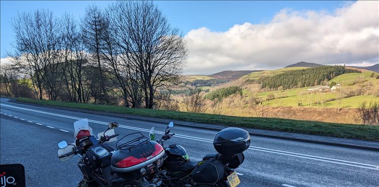 The 2 motorcycles outside in the shade, lovely hills in the sunlight beyond