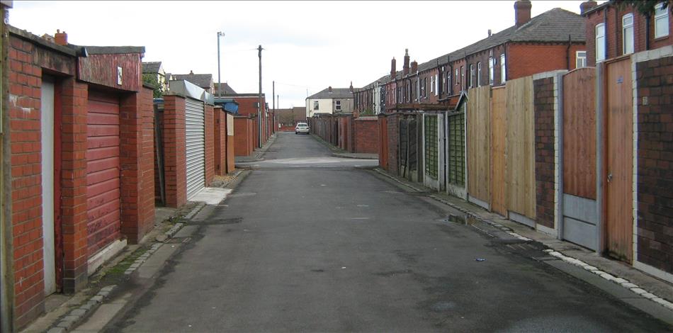 A back lane between terraced houses. There's garages and doorways and it's all a bit grim