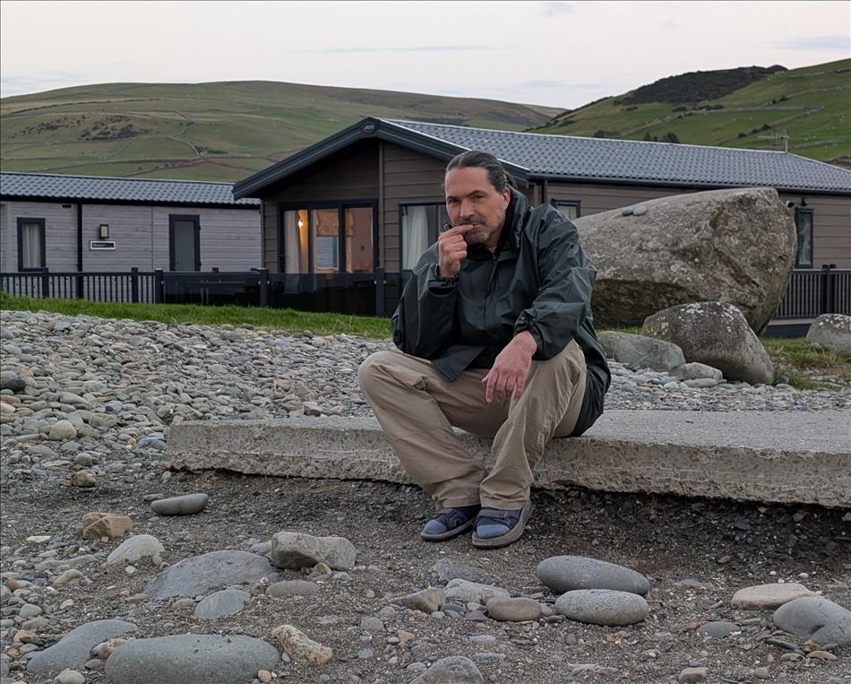 Ren sits on a concrete slab on a stony beach with hills and a few statics behind him, looking thoughtful