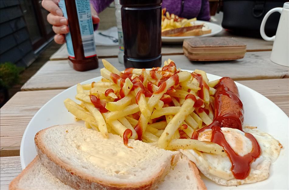 A plate with sausage, egg, chips and bread. there's sauce and salt and vineger on the table