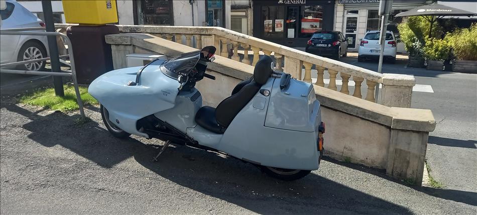 A peculiar motorcycle with the rider sat like in a car with feet forwards and one seat