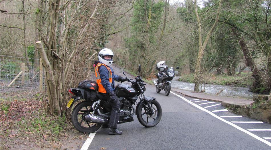 Sharon on her 125 and Ren's 125 behind on a narrow back lane with trees and a river